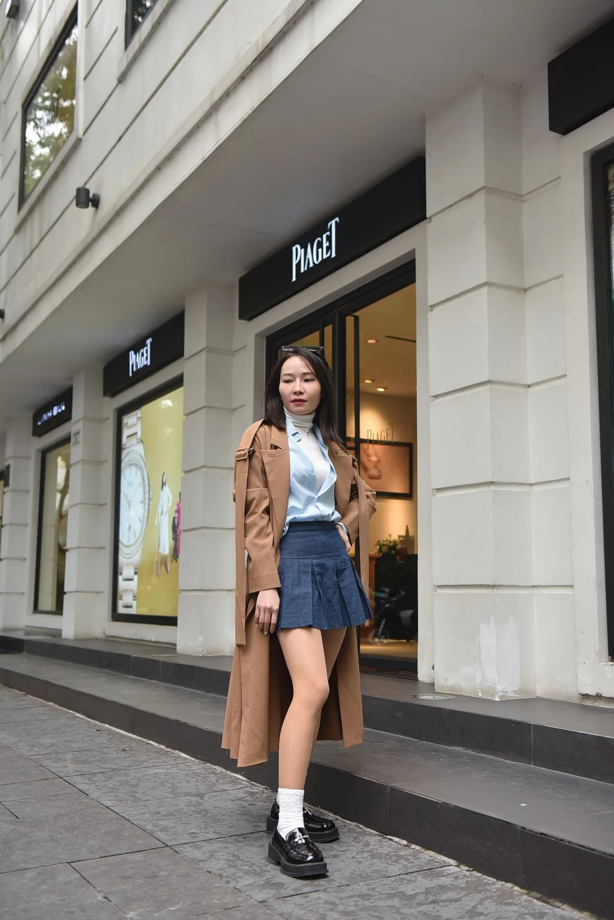 A woman in a long brown coat and short pleated skirt on a city pavement.