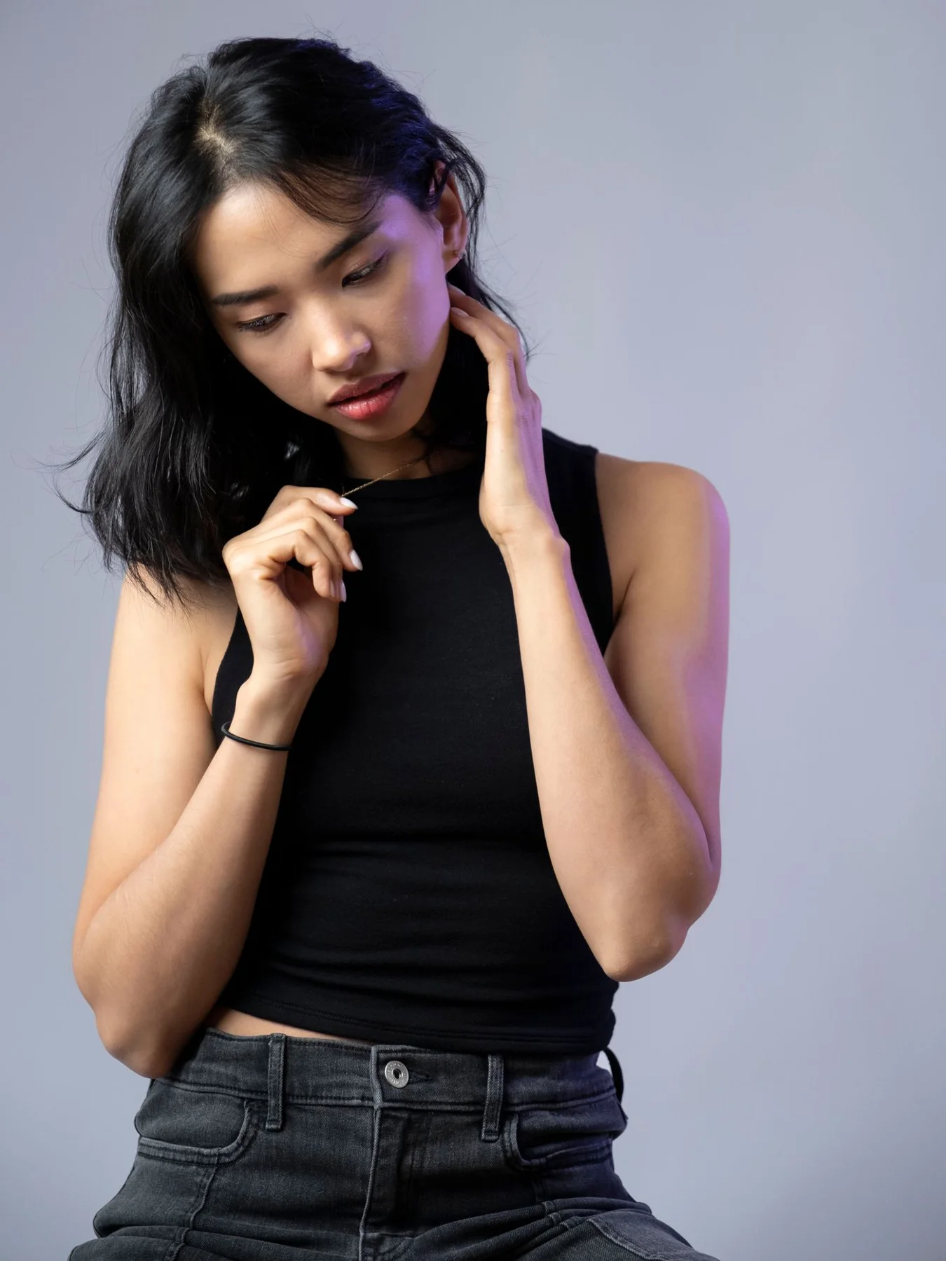 A woman wearing a dark sleeveless top and jeans against a grey background.