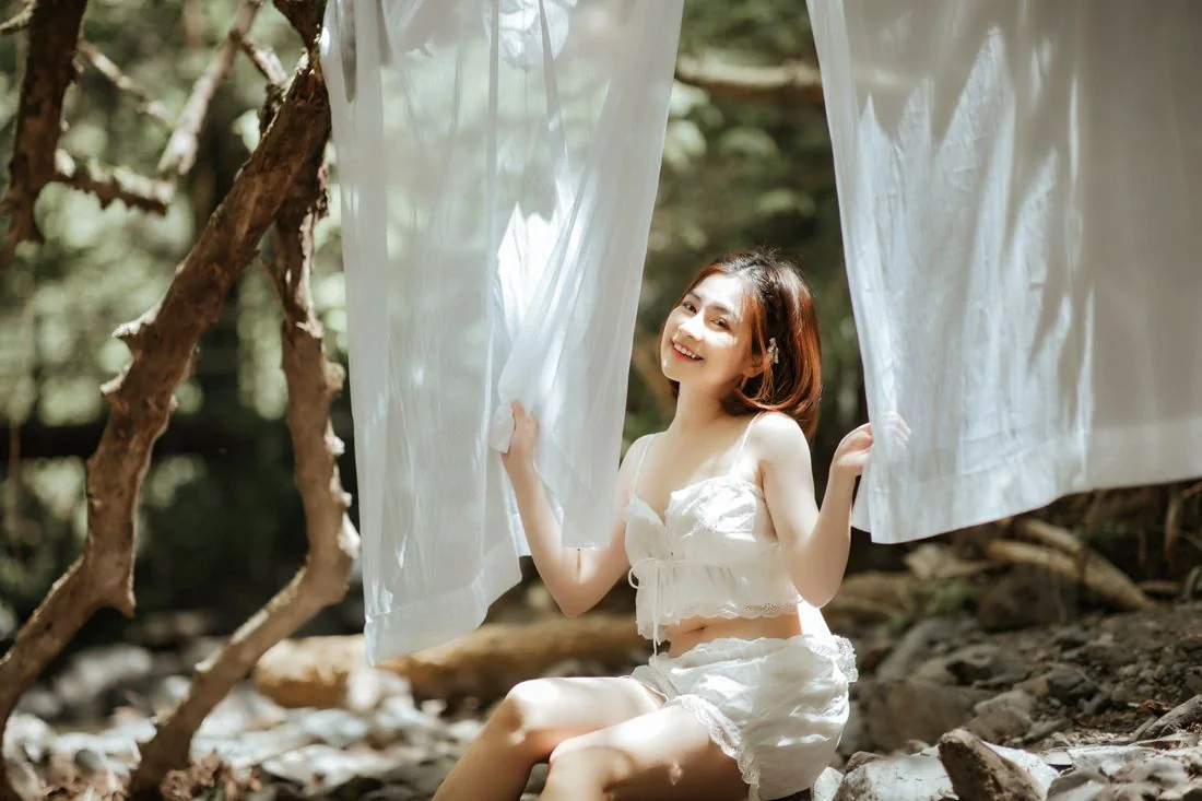 A woman in a pale outfit seated beside hanging pale fabric and trees.