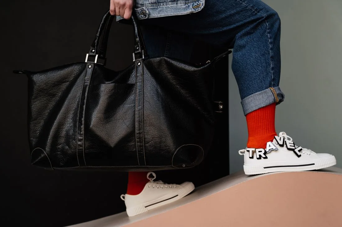 A dark travel bag beside pale sneakers, bright socks and rolled trouser hems.