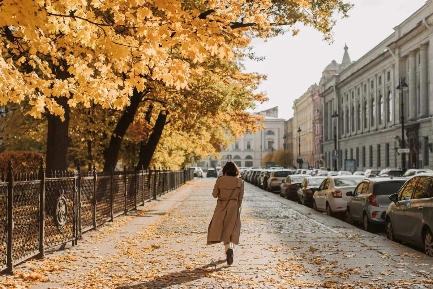 A woman in a long pale coat on a leaf-covered city pavement.