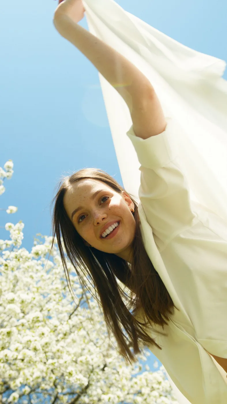 A woman in a flowing white garment beside flowering branches under a blue sky.